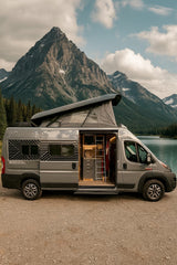Grey rv rental campervan with pop-top extended, side door open to its interior, parked by a lake & mountains.