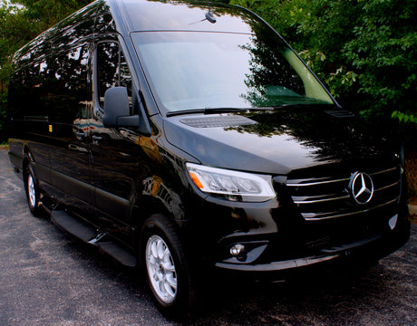 Black Mercedes-Benz Sprinter luxury van, an option for RV rental. Parked on asphalt, its shiny surface reflects green trees.