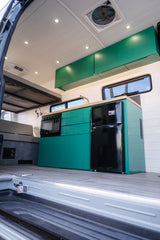 Modern RV rental interior: green kitchen cabinets, microwave, fridge, and sleeping area.