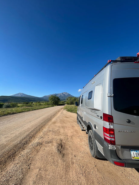 A silver Winnebago Revel RV rental is parked on a dirt road, facing away. Mountains rise under a blue sky, ideal for outdoor travel.