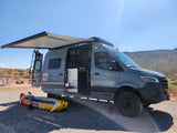 Grey camper van (RV rental) with awning & open doors, inflatable kayak ready. A sunny desert campsite awaits adventure.