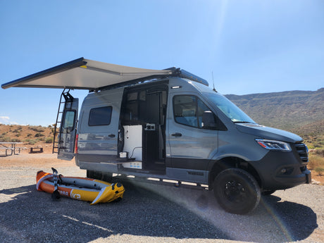 Grey camper van (RV rental) with awning & open doors, inflatable kayak ready. A sunny desert campsite awaits adventure.