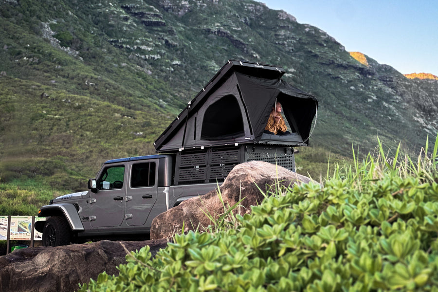 Adventure-ready RV rental! Jeep Gladiator with a rooftop tent parked on rocks near lush mountains for unique outdoor stays.