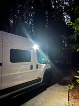 White RV rental van parked at night, illuminated by bright side lights against a dark forest background.