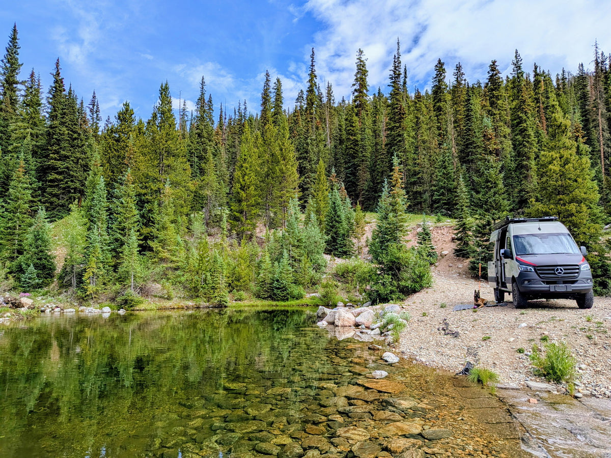 Grey camper van and dog next to a clear mountain lake with pine forests. Great spot for an RV rental escape.|Cream RV rental camper van parked on a dirt pull-off