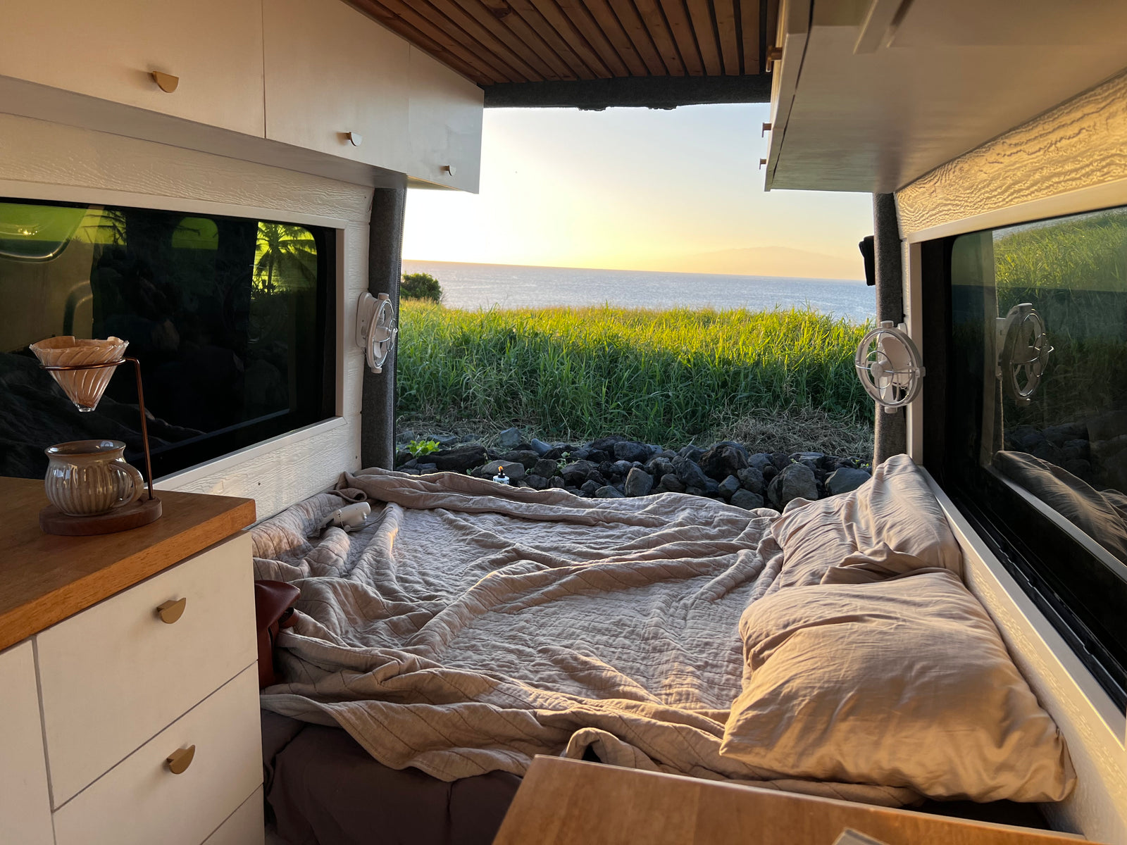 RV rental comfort: A cozy van interior with a bed, coffee setup, and stunning ocean sunset views from the window.