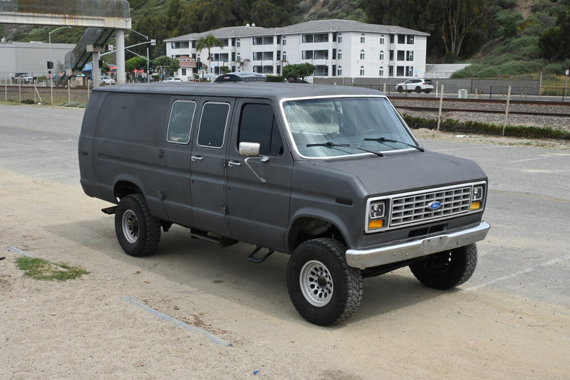 This matte gray lifted Ford Econoline van, possibly an RV rental, is parked on dirt near train tracks and buildings. — photo 8
