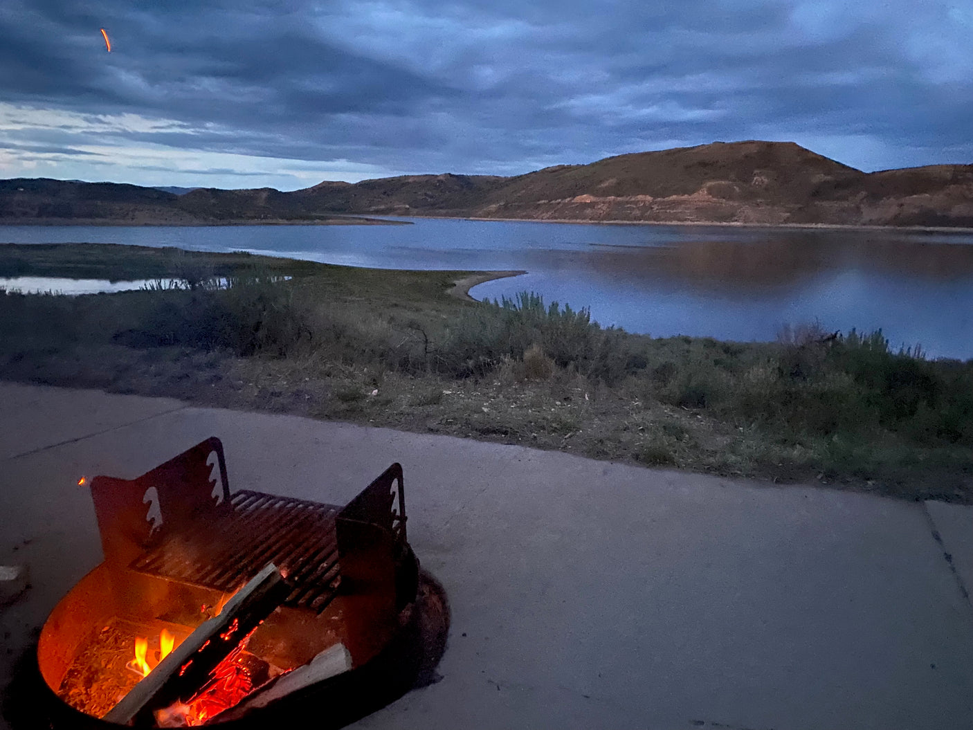 A cozy campfire burns by a serene lake and distant mountains under a cloudy dusk sky, ideal for an RV rental adventure.