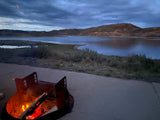 A cozy campfire burns by a serene lake and distant mountains under a cloudy dusk sky, ideal for an RV rental adventure.