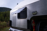A silver Kimbo truck camper, perfect for RV rental trips, shows its rugged design, open window with raindrops, and solar light.