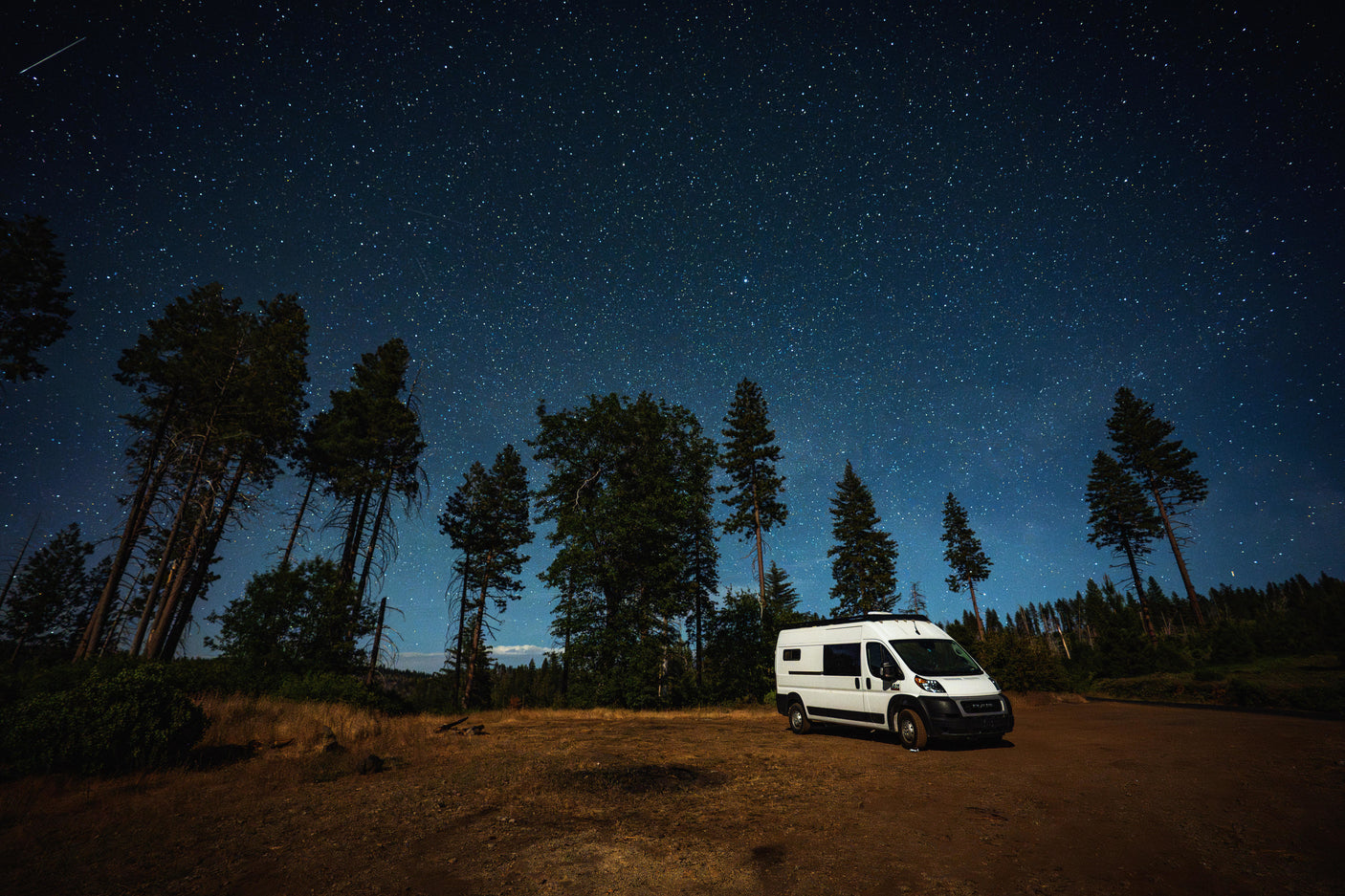 A white RV rental van is parked on a dirt path, nestled among tall trees, beneath a breathtaking, star-studded night sky.