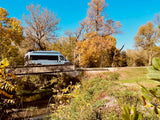 A light-colored rv rental camper van crosses a stone bridge over a tranquil creek in a vibrant autumn landscape.