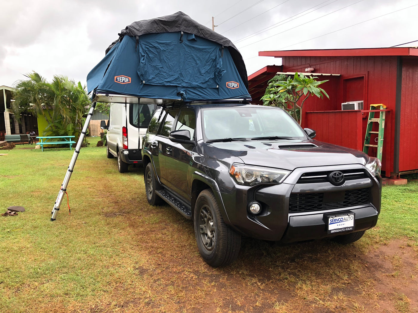 This RV rental features a grey SUV with a deployed blue Tepui rooftop tent and ladder, parked on grass near a white van. — photo 3