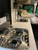 RV rental interior: A compact kitchen with a two-burner stove, sink, and a comfortable bed with a scenic window view.