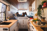 A man relaxes in a modern, organized RV rental with a cozy bed, well-equipped kitchen, and warm wood accents.