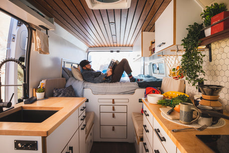 A man relaxes in a modern, organized RV rental with a cozy bed, well-equipped kitchen, and warm wood accents.