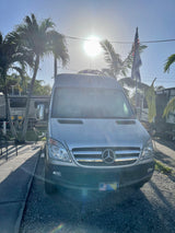 A silver Mercedes Sprinter van, a versatile RV rental, parked under sunny skies at a campground with palm trees and other RVs.