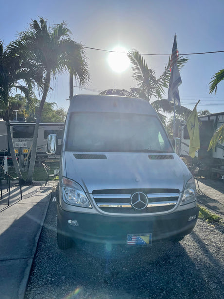 A silver Mercedes Sprinter van, a versatile RV rental, parked under sunny skies at a campground with palm trees and other RVs.