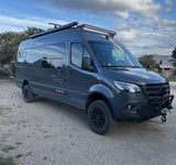 Dark gray Mercedes Sprinter rv rental camper van with off-road tires and light bars, parked on a gravel path under a cloudy sky.