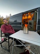 Happy man relaxing by his camper van rv rental. A portable fire pit glows on the table, setting cozy outdoor camping vibes.