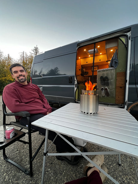 Happy man relaxing by his camper van rv rental. A portable fire pit glows on the table, setting cozy outdoor camping vibes.
