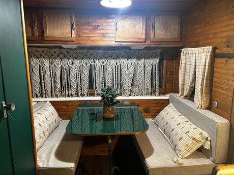 Inside a charming RV rental: Wood-paneled walls, a green dinette table, cozy seating, and macrame window decor.