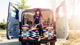 A woman sits in her open RV rental camper van, on a vibrant patterned blanket, looking out at the sunny outdoor scene.