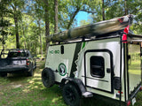 An RV rental camper with a kayak atop, parked with a truck in a wooded campsite, ready for adventure.