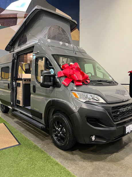 Grey Winnebago Ram camper van, featuring a pop-top and red bow, showcased for potential RV rental. Interior is visible.
