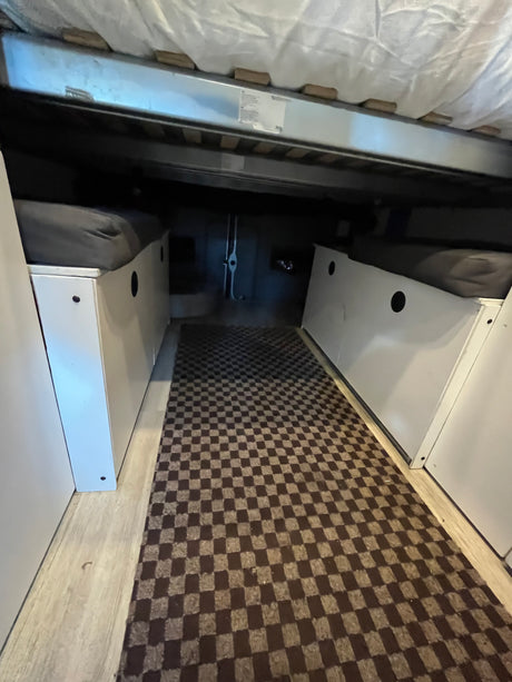 RV rental: Interior view of compact sleeping area with bed, side storage units, and a checkered aisle mat.