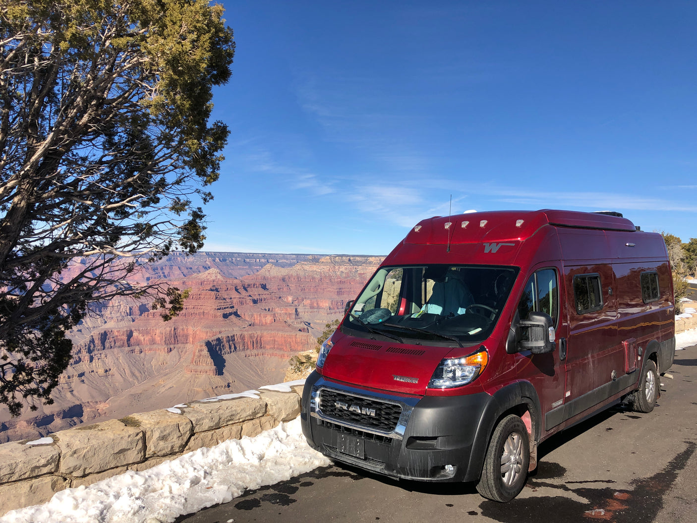 Red rv rental parked at the Grand Canyon viewpoint on a sunny day with snow, a tree, and canyon views.