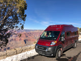 Red rv rental parked at the Grand Canyon viewpoint on a sunny day with snow, a tree, and canyon views.