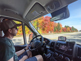 Driver in an RV rental on a scenic fall drive through Hiawatha National Forest, with phone navigation on display.