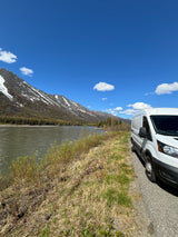 RV rental parked beside a scenic river with snowy mountains under a clear blue sky.