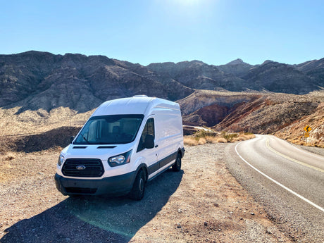 A white RV rental van, a Ford Transit, is parked beside a winding desert road with mountains under a bright blue sky.