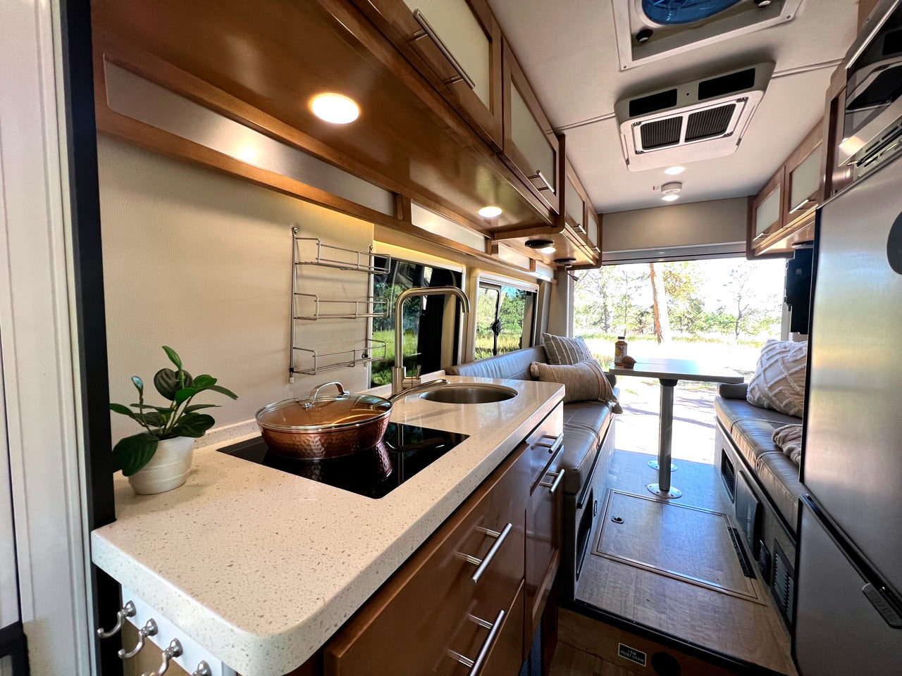 Modern RV rental interior showing a kitchen with a pot on the counter, leading to a bright dining area with a scenic view.