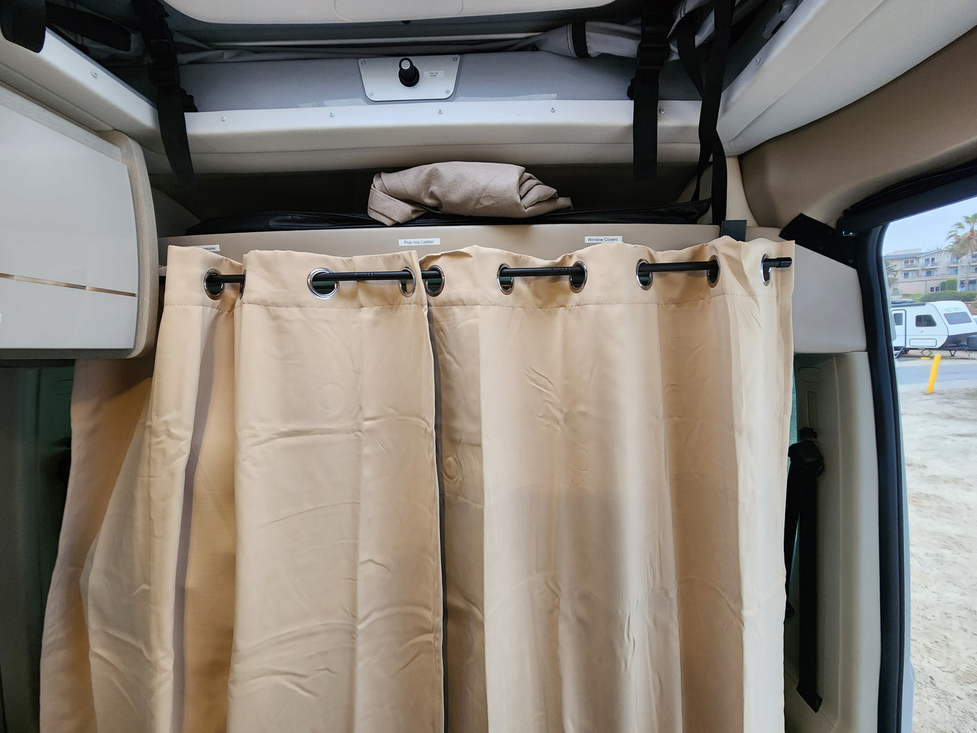Inside an RV rental, featuring beige privacy curtains, overhead storage labeled 'Pop-top Ladder' & 'Window Covers'.