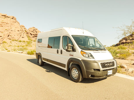 White Ram ProMaster RV rental van parked on a sunny desert road amidst rocky hills. Ideal for adventures!