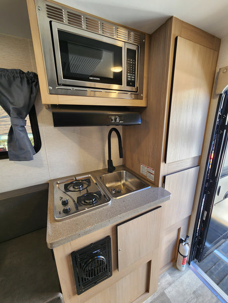 RV rental kitchen: Greystone microwave, 2-burner stovetop, sink, vent hood, and seating. Fire extinguisher visible.