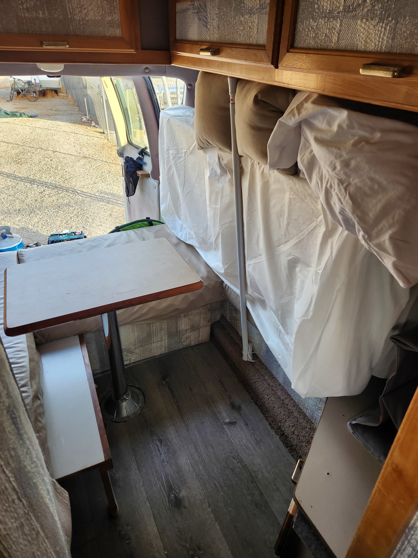 Inside a compact RV rental: a dinette with a bench and table, adjacent to a bed area, with an outside view.