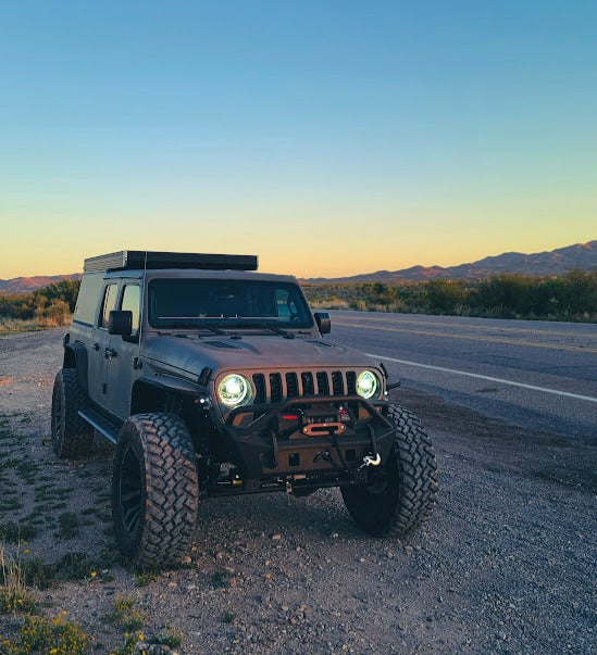 Gray modified Jeep Gladiator truck with a rooftop camper, parked on a desert road at dusk. A unique RV rental for off-road adventure.