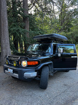 Black Toyota FJ Cruiser with a rooftop tent, parked among trees. Great rv rental for overlanding.