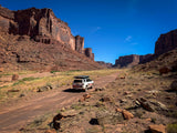 A white SUV with a roof tent, an RV rental, traverses a dusty road through towering red rock canyons under a blue sky.