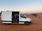 White RV rental parked in desert. Person and dog cozy in sleeping bag, open van door. Mesas under dawn/dusk sky.