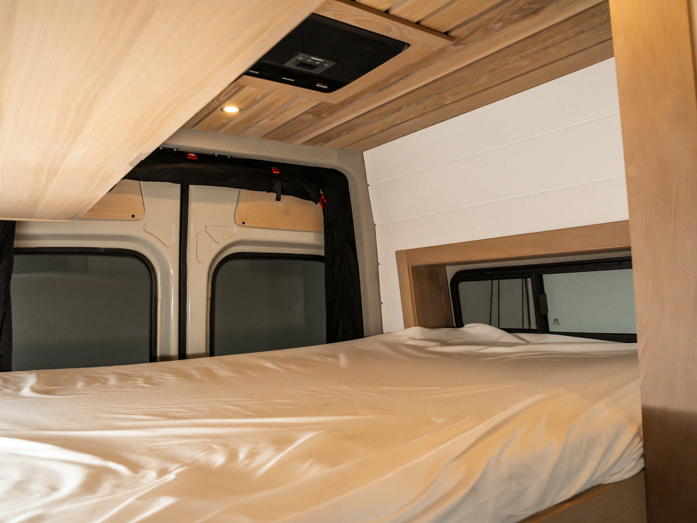 Cozy RV rental bed with a custom wooden ceiling, embedded light, and windows in a modern camper van sleeping area.