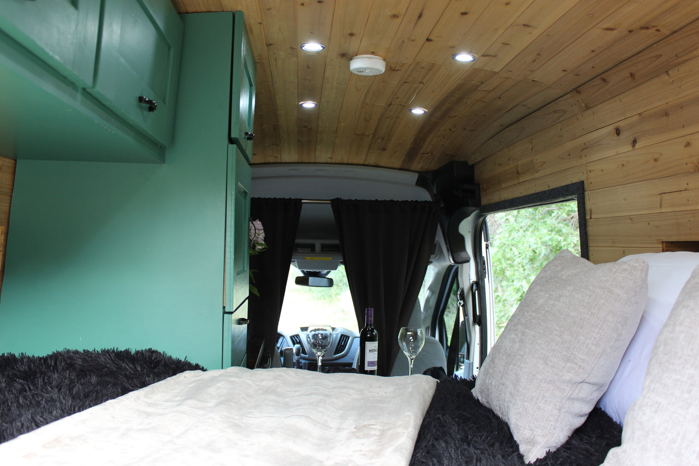Cozy rv rental camper van interior with a bed, green cabinets, wooden ceiling, and wine set up for a relaxing trip.