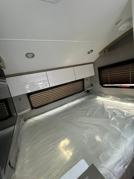 Bright RV rental bedroom showing a new, plastic-wrapped mattress, sleek overhead cabinets, and window blinds.