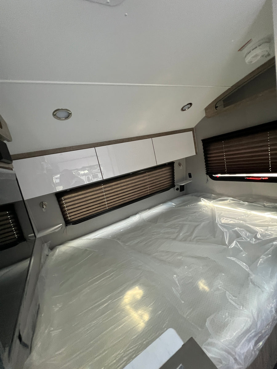 Bright RV rental bedroom showing a new, plastic-wrapped mattress, sleek overhead cabinets, and window blinds.
