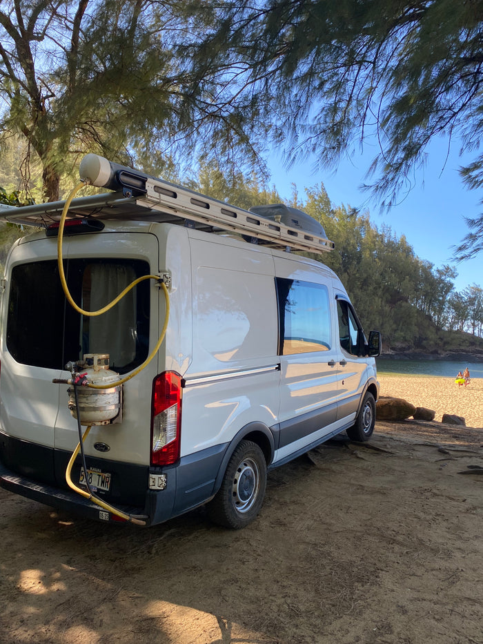 White Ford Transit rv rental camper van parked on a beach, equipped with a roof rack and outdoor shower.