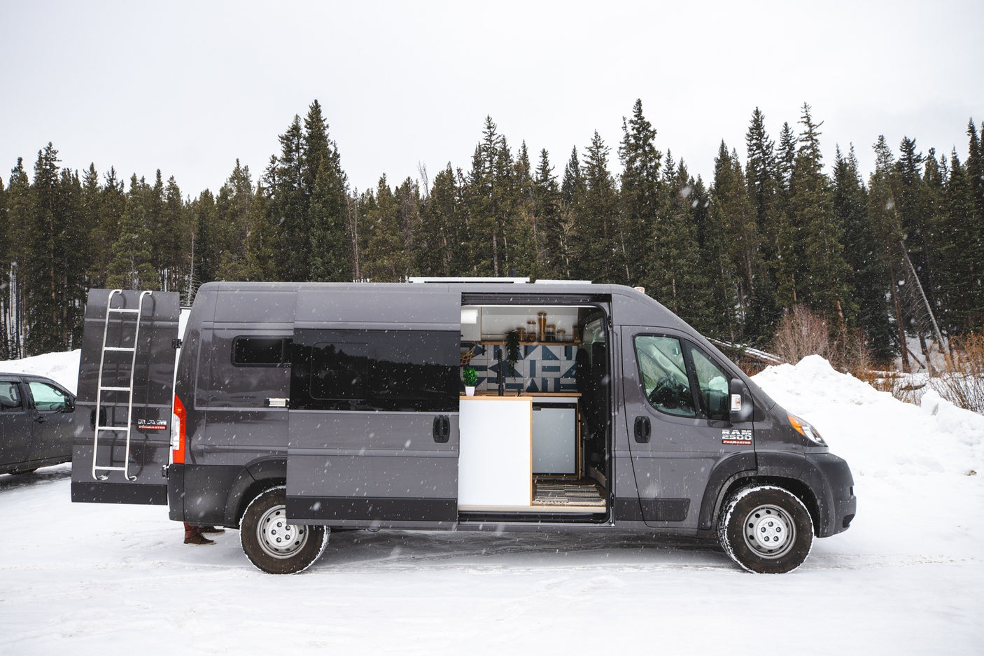 A grey Ram ProMaster rv rental, with open side and rear doors, parked in a snowy forest. Its custom interior is visible.